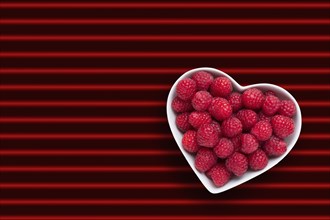 Raspberries in heart-shape bowl on red striped background