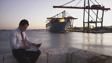 Hispanic businessman with laptop with container ship in background