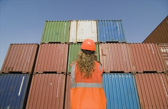 Woman wearing hard hat standing by containers in dock rear view