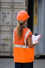 Female worker with clipboard checking containers in container terminal rear view