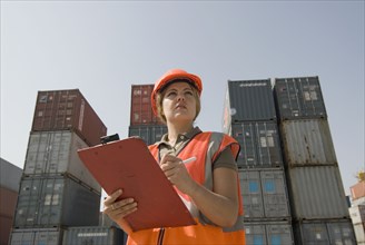 Woman wearing hard hat standing by shipping containers with clipboard