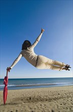 Woman jumping mid air on beach leaning on umbrella rear view