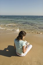 Young woman sitting on beach reading book rear view