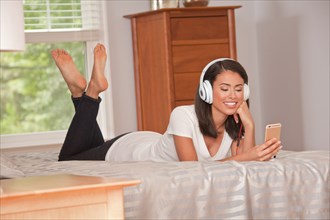Hispanic woman laying on bed listening to cell phone with headphones