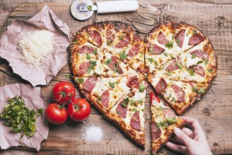 Hand pulling slice of heart-shaped pizza near ingredients on cutting board