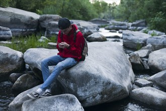 Caucasian man sitting on rock in river texting on cell phone