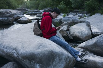 Caucasian man sitting on rock in river texting on cell phone