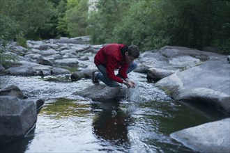 Caucasian man kneeling on rock in river cupping water with hands