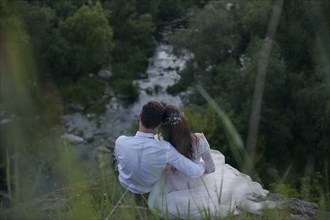 Caucasian bride and groom sitting on hill admiring river