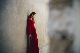Caucasian woman wearing red dress leaning on wall in alley