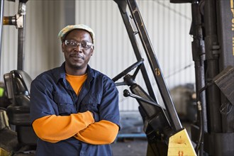 Smiling Black worker posing near forklift in factory