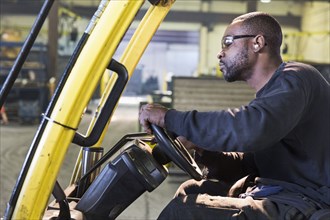 Black worker driving forklift in factory