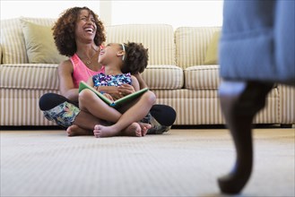 Laughing mixed race mother and daughter reading book on floor