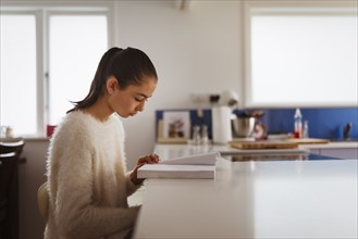 Mixed race girl reading book in kitchen