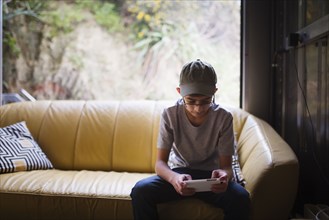 Mixed Race boy sitting on sofa near window texting on cell phone