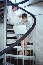 Mixed Race brother and sister sitting on spiral staircase using laptops
