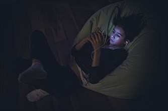 Mixed Race girl using cell phone laying on bean bag in dark