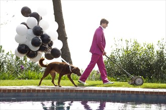 Caucasian man in pink suit mowing lawn being followed by dog