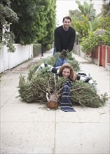 Caucasian couple dragging Christmas tree in alley