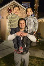 Hispanic man carrying sons on shoulders outside house decorated with string lights