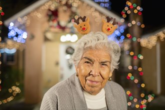 Older Hispanic woman smiling outside house decorated with string lights