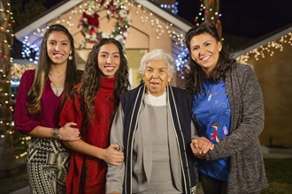 Three generations of Hispanic women smiling outside house decorated with string lights
