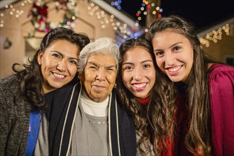 Three generations of Hispanic women standing outside house decorated with string lights