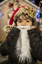 Hispanic girl wearing white beard and Santa hat outside house decorated with string lights
