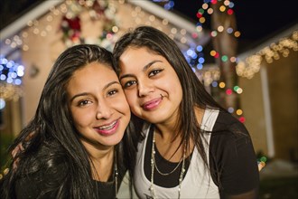 Hispanic sisters smiling outside house decorated with string lights