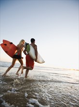 Couple running on beach carrying surfboards