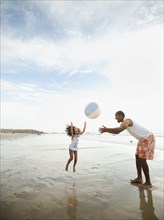 Black father throwing ball on beach with daughter