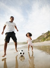Black father kicking soccer ball with daughter on beach