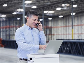 Caucasian businessman using laptop and cell phone in empty warehouse