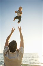 Caucasian father throwing baby son in air near ocean