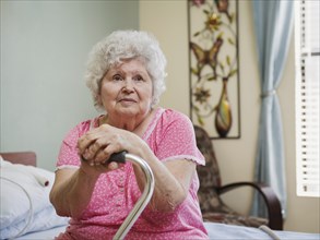 Serious Caucasian woman sitting on bed leaning on cane