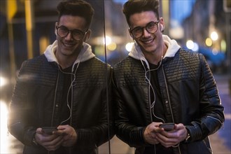Smiling Caucasian man leaning on wall listening to music on cell phone