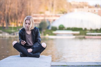 Caucasian woman sitting on wall near river texting on cell phone