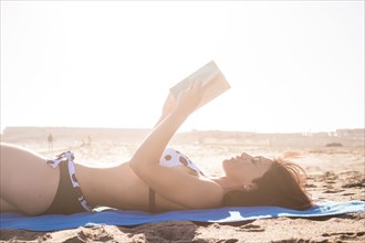 Caucasian woman laying on beach blanket reading book