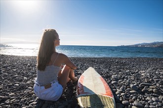 Caucasian woman sitting on beach near surfboard