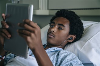 Black boy laying in hospital bed listening to digital tablet