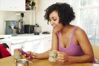 Mixed race woman using cell phone and drinking coffee in kitchen