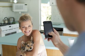 Man taking picture of wife holding dog in kitchen