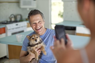 Woman taking picture of husband holding dog in kitchen