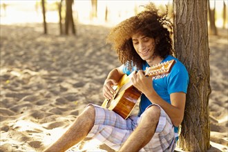 Mixed race teenager sitting in sand strumming guitar