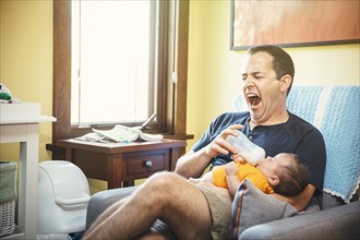 Yawning Caucasian father feeding baby boy in living room