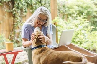 Caucasian woman using laptop petting dog on backyard patio