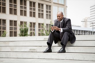 Black businessman sitting on staircase texting on cell phone outdoors