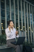 Businesswoman sitting on bench with suitcase talking on cell phone