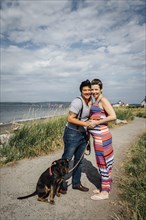 Portrait of pregnant lesbian couple with dog hugging at waterfront