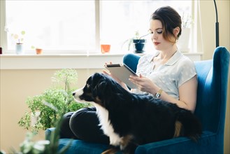 Woman sitting in armchair with dog using digital tablet